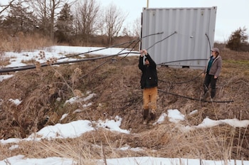 Un hombre y una mujer trabajan con cables cerca de un contenedor gris. El suelo tiene hierba seca y parches de nieve bajo un cielo nublado