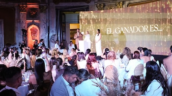 Vista general de un evento de gala. En el escenario, un presentador y dos personas ante una gran pantalla que anuncia 'EL GANADOR ES...'. La audiencia sentada en mesas redondas observa