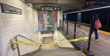 Una escalera de metro bloqueada con cinta amarilla de precaución, carteles de señalización de líneas y una persona caminando en el andén