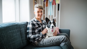 Una persona sonriente con cabello corto sentada en un sofá gris con un perro Corgi blanco y negro. El perro, con la lengua fuera, mira a cámara