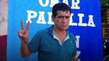 Un hombre con camiseta de rayas azules hace la señal de la V con los dedos frente a un cartel azul con su nombre