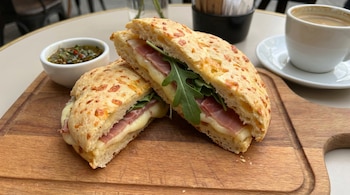 Vista cercana de un sándwich de pan de chipá cortado por la mitad, revelando capas de jamón, queso derretido y hojas de rúcula, sobre una tabla de madera.