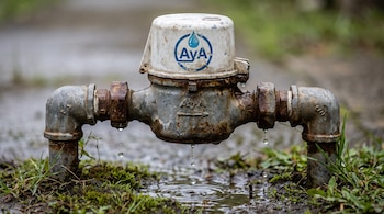 Medidor de agua metálico oxidado con tapa blanca que muestra el logo azul AyA, gotea sobre un charco pequeño de agua en el suelo con pasto.
