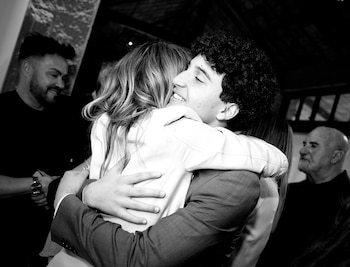 Fotografía en blanco y negro en la que un hombre joven de traje abraza con una sonrisa a una mujer rubia, entre otros invitados