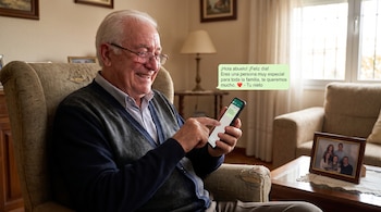 Hombre mayor con gafas sentado en un sillón, sonriendo y sosteniendo un teléfono móvil con un mensaje de WhatsApp. Una foto familiar está en una mesa.