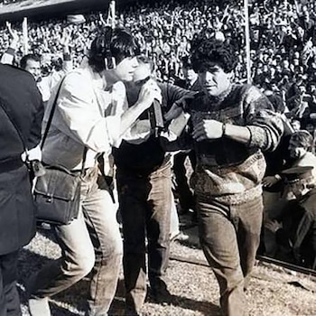 Diego Maradona camina sobre el césped junto a un reportero con micrófono y auriculares frente a una grada repleta de personas
