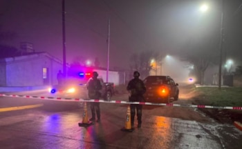 Carretera mojada de noche, dos agentes de seguridad con uniforme, cinta policial, dos conos, coches de policía con luces encendidas y niebla densa