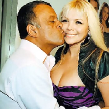 Un hombre de piel oscura besa la mejilla de una mujer rubia sonriente que viste un top morado y negro y una chaqueta oscura