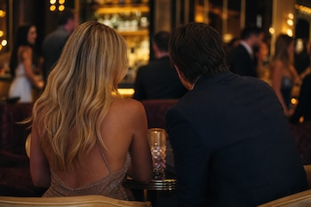 Una mujer de cabello rubio y un hombre de traje están de espaldas sentados a una mesa en un local con iluminación tenue.