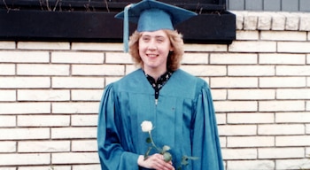 Mujer joven rubia con toga azul y birrete de graduación, sosteniendo una rosa blanca, de pie frente a una pared de ladrillos clara