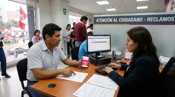 Hombre consulta a mujer sentados en un escritorio con computadoras, documentos, teléfono celular que muestra Indecopi y bandera de Perú en el fondo.