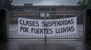 Una pancarta con el texto CLASES SUSPENDIDAS POR FUERTES LLUVIAS está colgada de un portón metálico bajo la lluvia frente a un edificio.