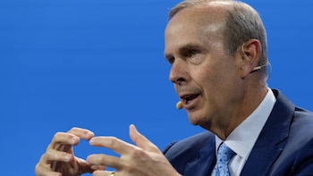 Primer plano de Mike Wirth, un hombre con traje azul y corbata clara, gesticulando con las manos. Lleva un micrófono cerca de la boca. Fondo azul liso