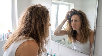 Mujer con cabello castaño largo y despeinado frente a un espejo en un baño, con el reflejo de su rostro que muestra preocupación mientras toca su cabeza.