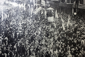 Fotografía en blanco y negro de una multitud de personas en una calle con banderas y edificios en los costados