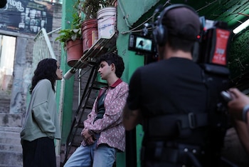 Un camarógrafo filma a dos actores, un hombre y una mujer, conversando en una calle urbana con paredes verdes y plantas en macetas