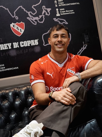 Un hombre con camiseta deportiva roja posa sentado en un sofá de cuero negro frente a una pared decorada con logotipos