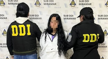 Dos agentes policiales de espaldas custodian a una mujer con el rostro pixelado frente a un fondo con inscripciones oficiales