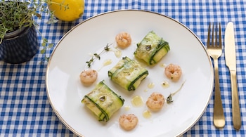 Plato con tres rollos de calabacín, siete gambas cocidas, hierbas, aceite. Un limón, planta en maceta, cubiertos dorados y mantel a cuadros azul y blanco.