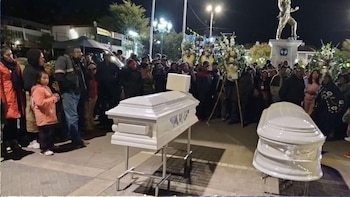Fotografía nocturna de un funeral al aire libre con dos ataúdes blancos sobre pedestales, rodeados por una multitud de personas y arreglos florales