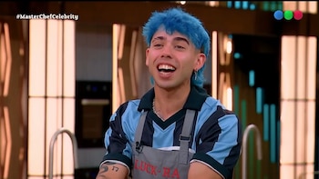Un joven con el cabello teñido de azul y un delantal gris sonríe dentro de un estudio de televisión