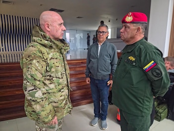 Tres hombres en un interior. Uno con uniforme de camuflaje, otro con ropa civil, y el tercero con uniforme militar verde, boina roja y bandera de Venezuela