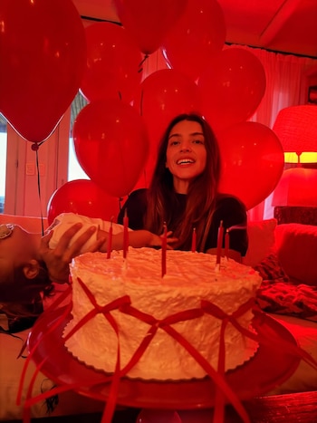 Calu Rivero sonriendo detrás de una torta blanca con cintas rojas y velas, rodeada de globos rojos, con un bebé en brazos y Aito de la Rua cerca