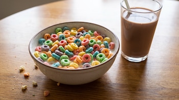 Un tazón cerámico con cereales multicolores y leche junto a un vaso con chocolatada y una pajilla sobre una mesa de madera.