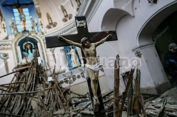 Un crucifijo grande de madera y figura de Cristo en el centro de escombros de madera y mampostería, dentro de una iglesia con estatuas y un arco al fondo