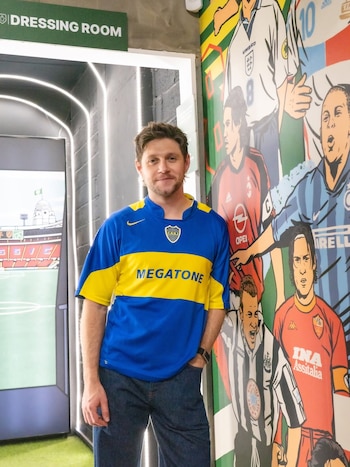 El cantante Niall Horan sonríe mientras posa de pie, vistiendo una camiseta azul y amarilla de Boca Juniors, con un mural de figuras del fútbol detrás