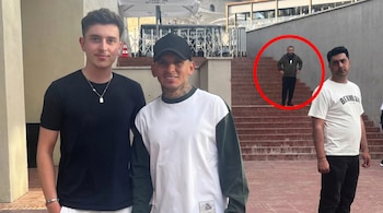 Dos jóvenes sonríen en primer plano, con un tercer hombre cerca. Detrás, un hombre barbudo con chaqueta verde y manos en caderas en escaleras de ladrillo observa.