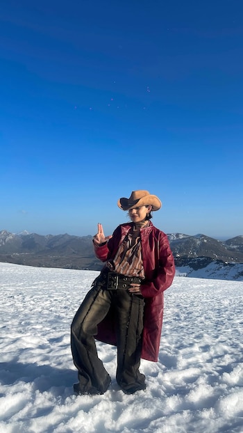 Dagna Mata, vista de cuerpo completo, posa en un paisaje nevado con montañas al fondo y cielo azul. Viste sombrero vaquero, chaqueta roja, pantalón oscuro y hace un signo de paz