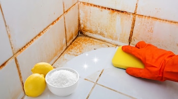 Azulejos blancos de baño con suciedad y sarro en las juntas, una mano con guante naranja y esponja amarilla limpia una sección, junto a dos limones y un tazón con sal.