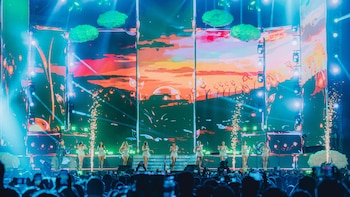 Escenario con pantallas LED que muestran un paisaje digital, luces de concierto, fuegos artificiales, grupo musical femenino, un cantante y público con teléfonos