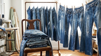 Pantalones jeans apilados en una silla de madera y colgados en un tendedero con pinzas y un perchero metálico; se ven una máquina de coser y rollos de tela.