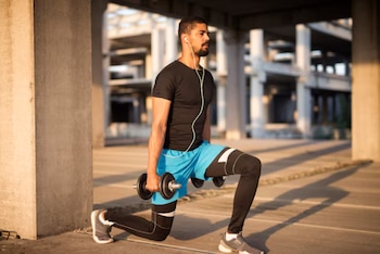 Hombre con camiseta negra, shorts azules y mallas negras realiza una zancada con dos mancuernas. Lleva auriculares y zapatillas deportivas