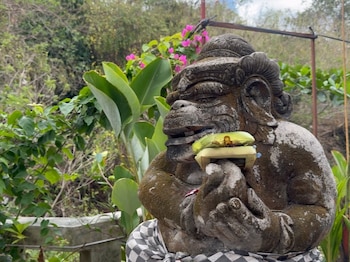 Estatua de piedra con tela a cuadros que sostiene una pequeña caja de ofrenda frente a follaje verde y flores rosadas