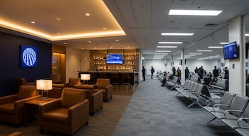 Sala VIP con logo de United Airlines, sillones de cuero y barra; sala de espera con sillas de plástico, pantalla de vuelos y personas en aeropuerto.