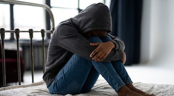 Persona con piel morena, sudadera gris con capucha, jeans azules, sentada en una cama con las rodillas recogidas, el rostro oculto. Fondo blanco y oscuro.