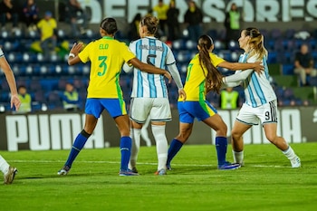 Cuatro jugadoras de fútbol femenino en el campo. Dos de Brasil con camisetas amarillas y shorts azules, dos de Argentina con camisetas celestes y blancas a rayas
