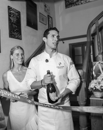 Hombre en chaqueta de chef, Rodrigo Fernandini, sonríe sosteniendo una botella de champagne detrás de un listón con 'FERNANDINI'. Mujer rubia sonríe a su izquierda