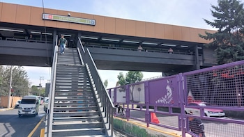 Vista exterior de la estación Metro Universidad con personas subiendo y bajando escaleras y una pasarela superior. Se ven vehículos en la calle y una valla morada