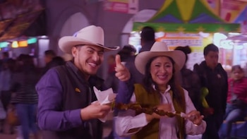 Un hombre y una mujer sonrientes con sombreros de cowboy. El hombre sujeta un papel blanco. La mujer sostiene una brocheta de comida y levanta el pulgar