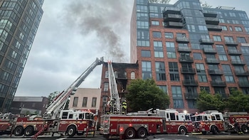 Varios camiones de bomberos rojos con escaleras extendidas combaten un incendio en un edificio de ladrillos, del cual emana humo denso, en un entorno urbano