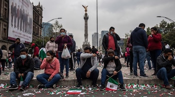 Grupo de personas con mascarillas faciales, sentadas y de pie. Confeti tricolor cubre el suelo. Monumento El Ángel de la Independencia al fondo. Una bandera de México pequeña.