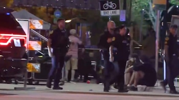 Policías en uniforme, una patrulla y varias personas, algunas sentadas, en una calle oscura de noche, posiblemente acordonada tras un incidente