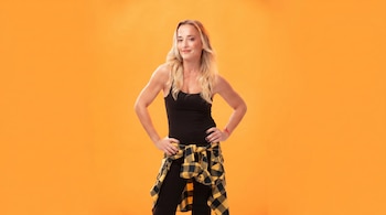 Mujer rubia con top negro, pantalones negros y camisa de cuadros amarilla y negra en la cintura, posa sonriendo con manos en las caderas sobre fondo naranja.