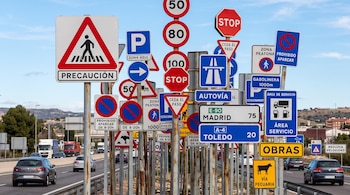 Diversas señales de tráfico de velocidad, prohibición e indicación colocadas en postes metálicos junto a una carretera con automóviles.