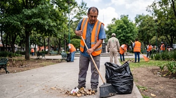 Un hombre con chaleco de seguridad naranja barre hojas secas con una escoba en el sendero de un parque junto a una bolsa de basura negra.