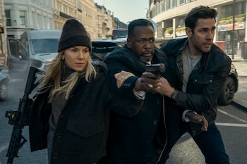 Una mujer rubia con gorro oscuro y rifle, un hombre afroamericano con pistola y un hombre blanco corren en una calle. Edificios y vehículos de fondo